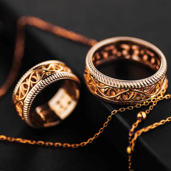 Du auksiniai žiedai su ornamentiniais raštais, padėti ant juodo paviršiaus su grandinėle