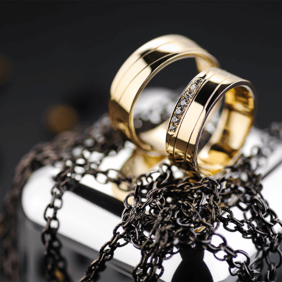 Dvi auksinės žiedų juostos su deimantais, padėtos ant metalinio paviršiaus su grandinėle